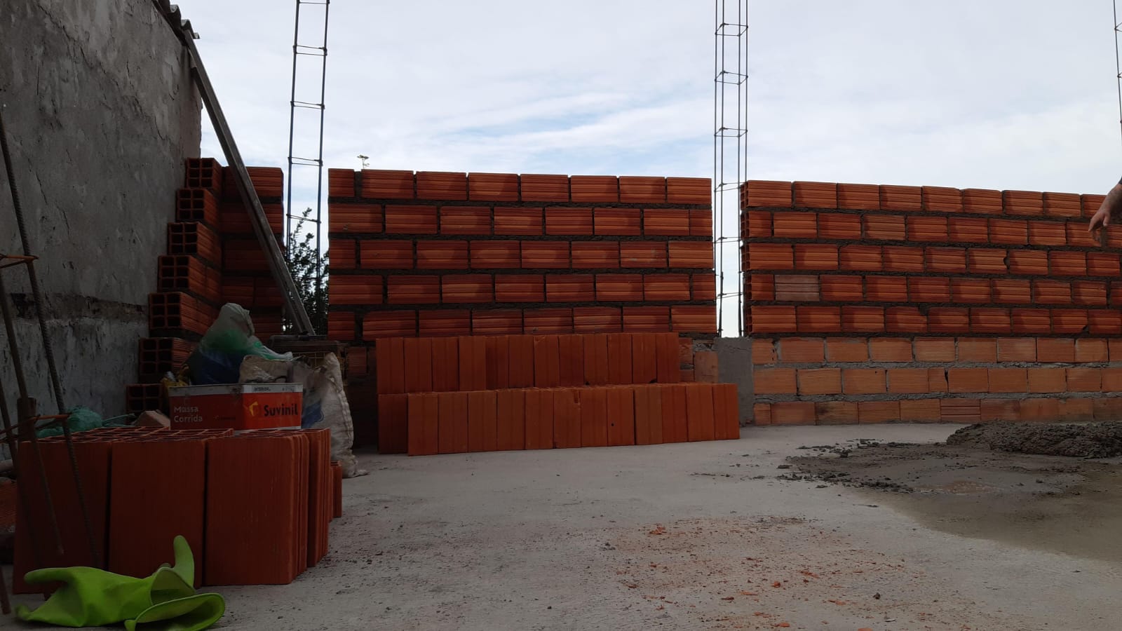 obra em construção com paredes de tijolos vermelhos sendo levantadas, pilhas de tijolos no chão, caixa de massa corrida e luva verde